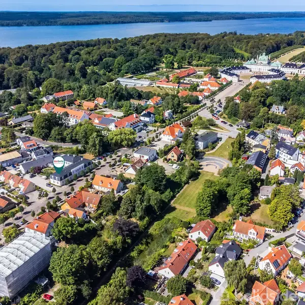 Byområde med røde tage spredt mellem grønne træer nær en sø. Området er omgivet af skov, og i baggrunden ses et slot.