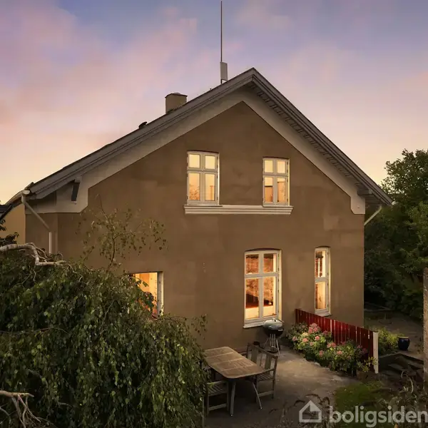 Huset er to etager højt med oplyste vinduer, der antyder indendørs aktivitet. Det står i en have med grønne træer og buske omkring, mens en udendørs terrasse er synlig.