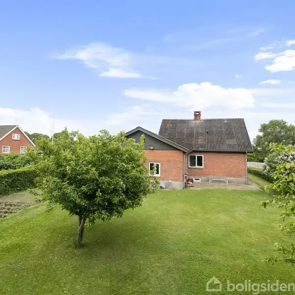 Et rødt murstenshus står stille i en frodig have med grøn græsplæne og et enkelt træ. Andre huse ses i baggrunden under en blå himmel med få skyer.