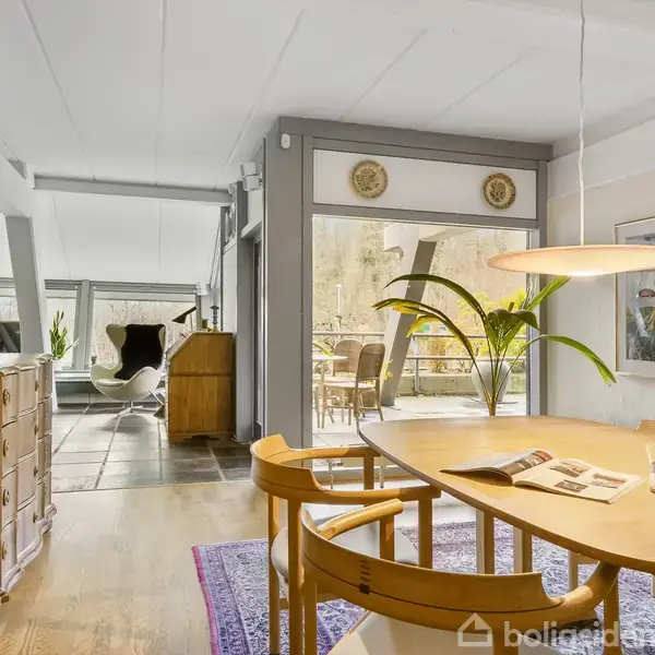 Et træskab står i en entré med brune klinkegulve. En spejlramme og to lamper er på væggen. Døren fører til en stue med lamper og planter.