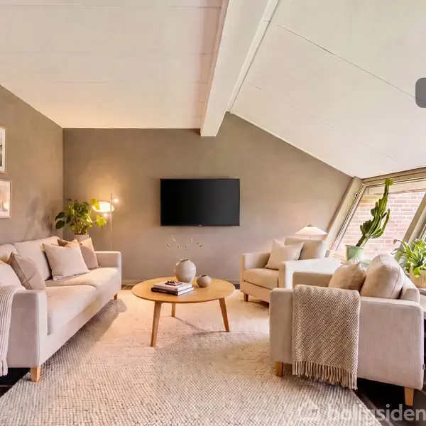 Stue med sofaer og en lænestol arrangeret omkring et rundt sofabord. En fladskærm er vægmonteret. Store skråvinduer og planter lysner rummet. Tekst: "Visualisering".