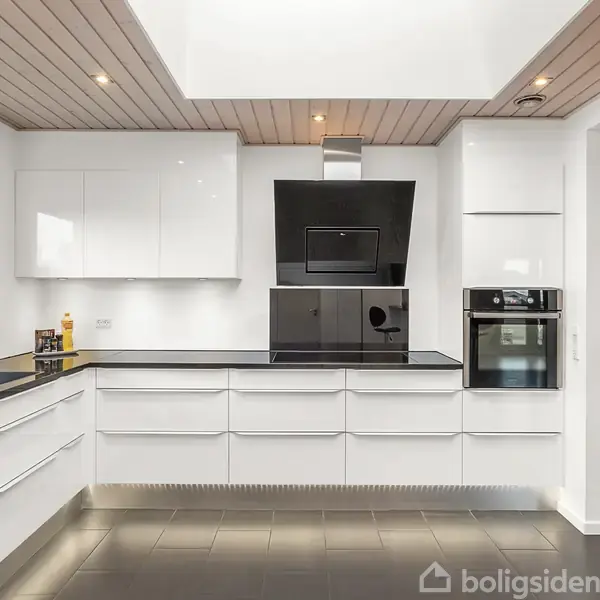 Køkken med hvide, moderne skabe, indbygget ovn i væggen og vask på venstre side. Rummet har fliser på gulvet og træloft, med et vindue der lukker naturligt lys ind.