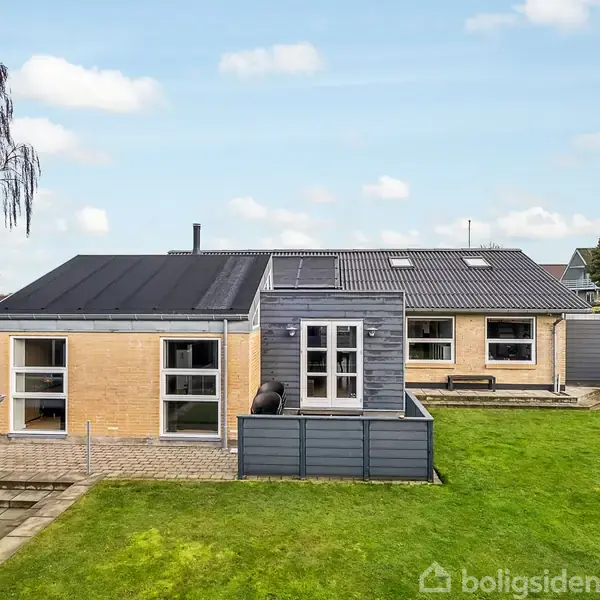 En gul murstenshus med sorte tage ligger midt i en grøn græsplæne. Solen skinner, og himlen er blå med få skyer. En terrasse ses til venstre.