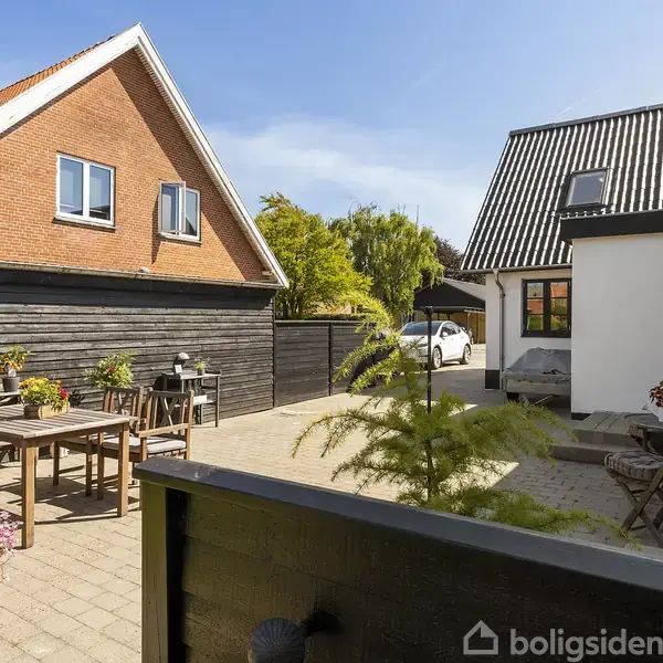 En gårdhave med bord og stole står fredeligt på flisebelagt område. Rundt om er der planter i krukker. Baggrund viser to huse med skråt tag og en klar blå himmel.