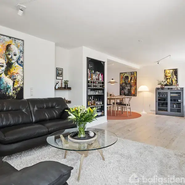 En stue med sort lædersofa og rundt glasbord med blomster. Farverige malerier hænger på væggene. Ved siden af er der en spiseplads med hylder og stort vindue.