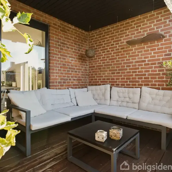 En komfortabel hjørnesofa med hvide puder står på en træterrasse. Sofaen omgives af murstensvægge og grønne planter, og et lille sofabord er placeret foran den.