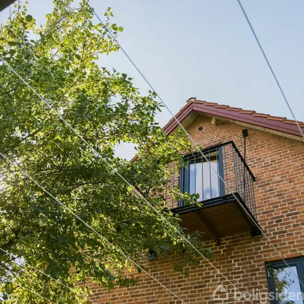 Et murstenshus har en lille balkon på første sal. Sollys skinner ind gennem bladene på et nærliggende træ, og der er ledninger i luften foran huset.