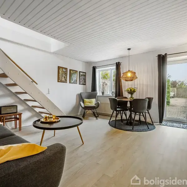 Stue med en trappe til venstre og en grå sofa med gule puder. En rund spisebord med stole står ved siden af et stort vindue med udsigt til en have.