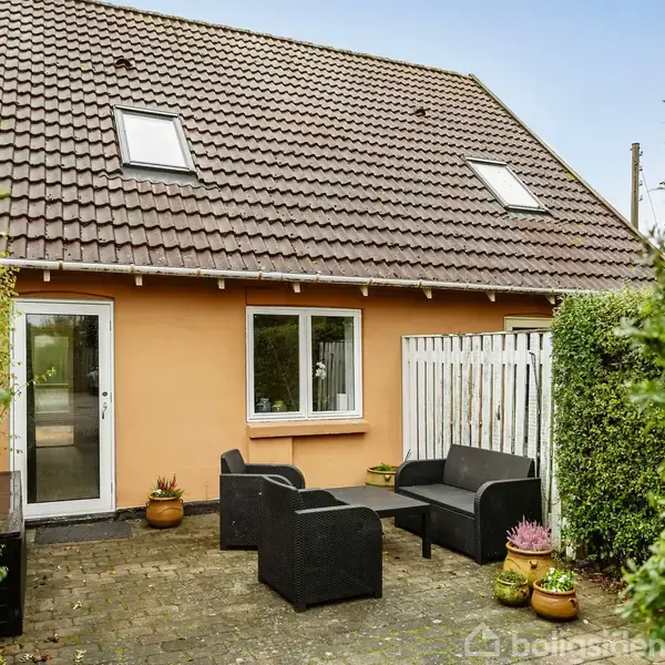 Et hus med rødt tegltag har en terrasse med sorte havemøbler. Omgivet af grønne hække, står der krukker med planter ved siden af døren og vinduer.