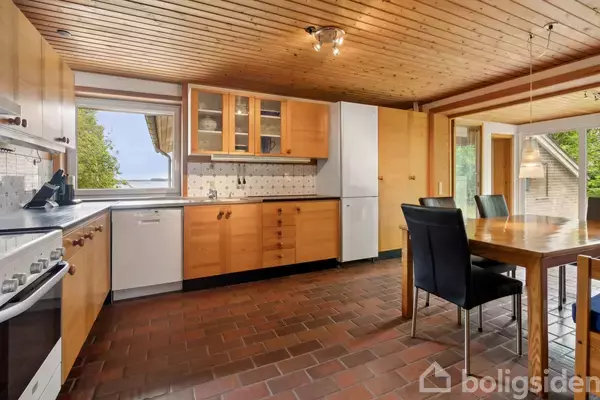 Køkken med træloft og gulvfliser, køkkenbord med skabe, ovn og køleskab. Spisebord med sorte stole står centralt. Udsigt til træer gennem vinduet.