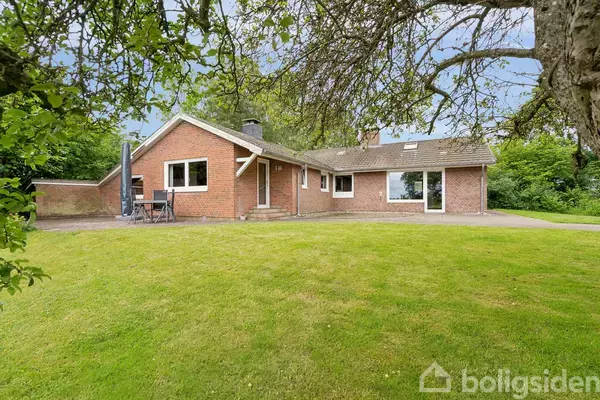 En rød murstensvilla med store vinduer, omgivet af en grøn have med et stort græsareal, står under en blå himmel med frodigt løv, der giver en idyllisk fornemmelse.