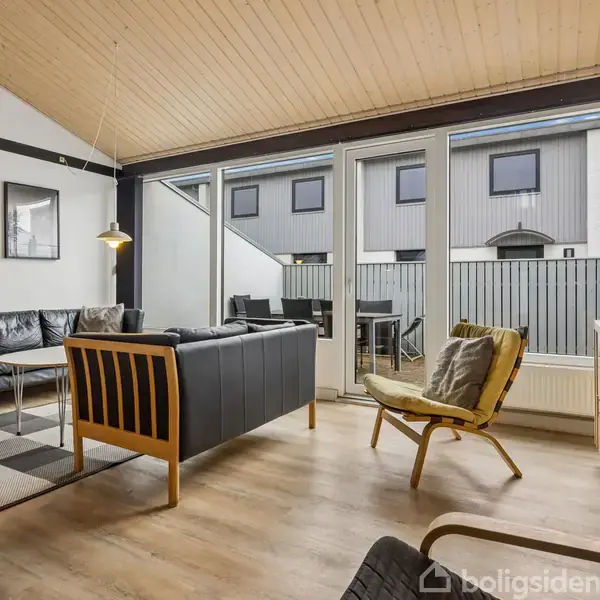 Stue med sorte lædersofaer og stole, sofabord, og gulvtæppe. Store vinduer med udsigt til en balkon. Træloft og moderne belysning. Ingen tekst til stede.