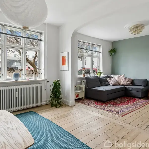Stue med en grå hjørnesofa placeret på et orientalsk tæppe, omgivet af lyse trægulve og hvide vægge. Store vinduer giver naturligt lys, og rummet indeholder planter og en papirlampe.