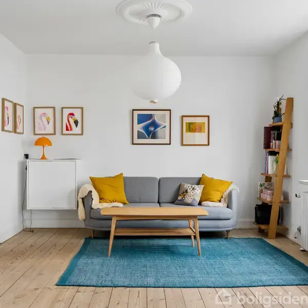 Grå sofa står på blå tæppe i lys stue; dekoreret med gule puder, vægkunst, orange lampe og træreol ved vindue. Hvidt loft med stor rund lampe.
