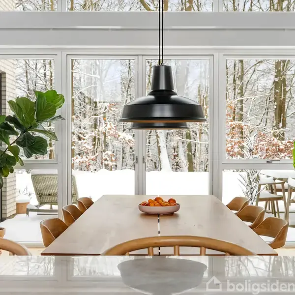 En spisestue med et langt bord, omgivet af polstrede stole, står midt i rummet. En stor, sort lampe hænger over bordet. Store vinduer viser en sneklædt have.