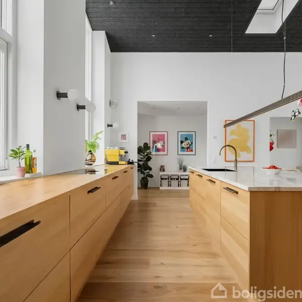 Et moderne køkken med træskabe og en hvid køkkenø placeret centralt. Store vinduer lyser rummet op, mens potteplanter og kunst på væggen skaber en hyggelig atmosfære.