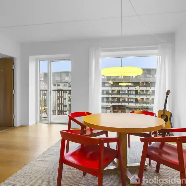 Rundt træbord med røde stole står i spisestue. En guitar læner sig op ad væggen ved siden af en reol fyldt med bøger. Udsigt til lejlighedskompleks gennem vinduer.