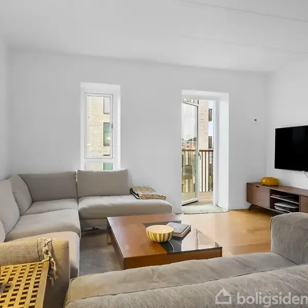 Sofa i hjørne står roligt i stue med grå vægge og træbord. Fjernsyn hænger på væggen ved balkon med glasdør. Lys lampe og små pyntegenstande på bordet.