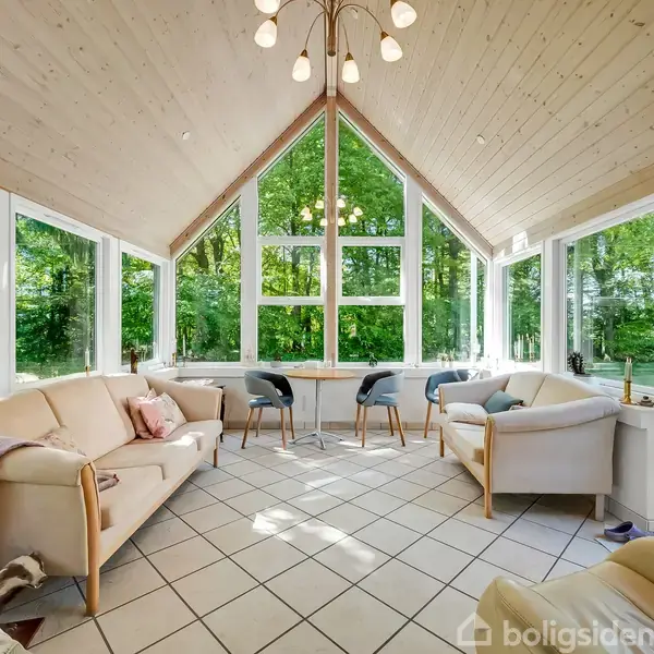 Lys stue med beige sofaer og stole, placeret på flisegulv. Store vinduer omgiver rummet og viser en frodig, grøn skov udenfor. Elegant lysekrone hænger fra træloftet.