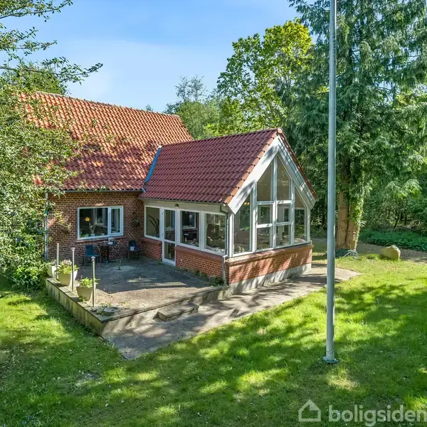 Et murstenshus med rødt tegltag og stor glaseret udestue ligger i en grøn have med træer og buske omkring. En flagstang står ved siden af huset på en græsplæne.