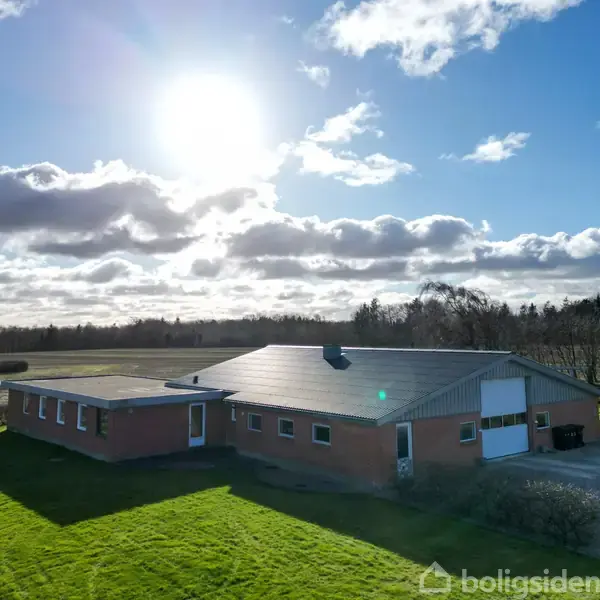 En enetages, rød murstensbygning står på en stor, åben græsplæne under en klar, solrig himmel med spredte skyer. Øde landskab og træer omgiver huset i baggrunden.