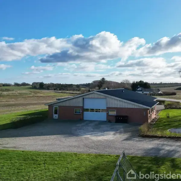 En stor ladebygning står stille på en grusbelagt gårdsplads omgivet af frodige marker og grønne arealer under en klar blå himmel med spredte skyer.