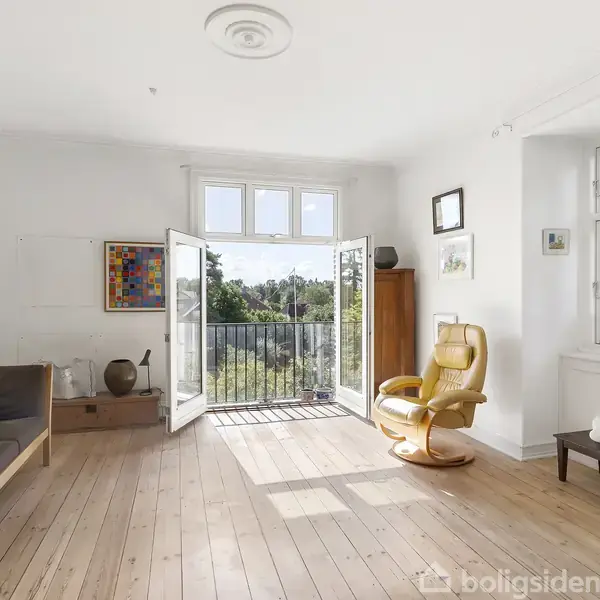 Stue med en åben dør til en balkon, en lænestol og sofa. Væggene er dekoreret med billeder, og der er gulvbrædder. Udsigt til en grøn have gennem vinduet.
