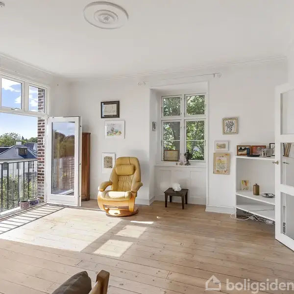 En gul lænestol står i et lyst stuehjørne med åbne døre til en balkon. Rummet har trægulv, hvide vægge, og hylder med bøger og billeder.