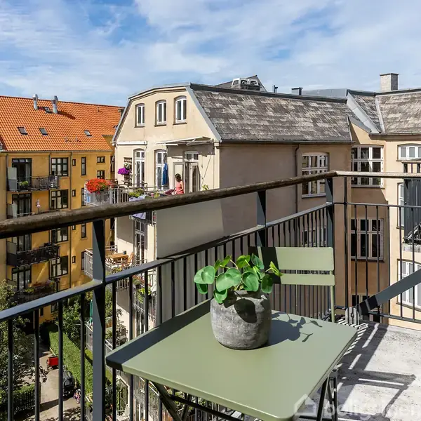 En lille balkon med et grønt bord og en potteplante, der står stille. Udsigt over tætte byhuse med røde tage og blomster i vinduerne under en blå himmel.