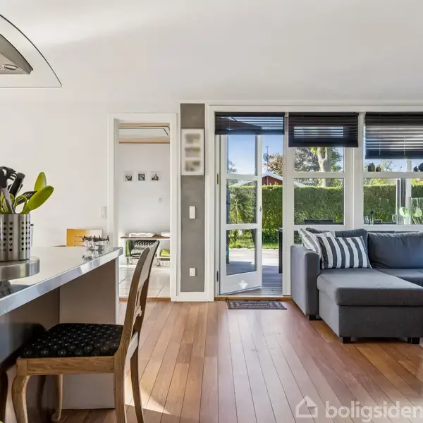 Køkken og stue kombineret; køkkenbord med køkkenredskaber til venstre; grå hjørnesofa til højre; store vinduer med udsigt til en have; trægulve skaber en lys og åben atmosfære.