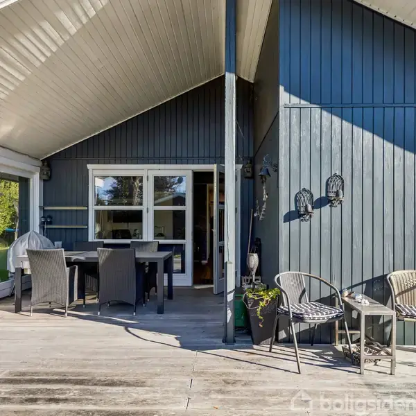 Et moderne sommerhus med træbeklædning, hvor en overdækket terrasse har spisebord og stole. Det omkringliggende område viser en have med grønne planter og en udendørs grill.