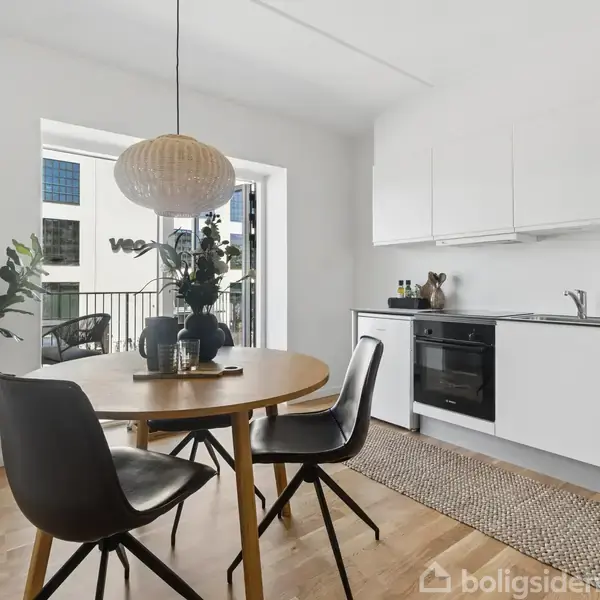 Rundt spisebord med sorte stole står i et lyst køkken. Ved siden af ses en åben altandør med udsigt til bygninger udenfor. Grønne planter dekorerer rummet.