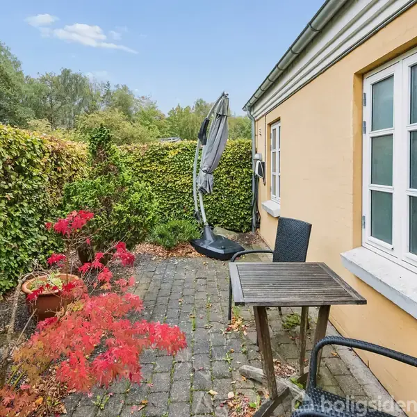 En træterrasse med to stole og et bord står langs en gul murstensbygning. En parasol er foldet sammen ved siden af. Omkring er der grønne buske og røde planter.