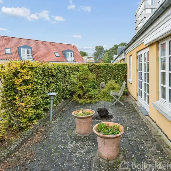 Murstensgårdhave med to krukker på flisebelægning. Huset har lyse gule vægge og store vinduer. En stol står i hjørnet. En hæk omgiver området, og der ses rødt tag i baggrunden.