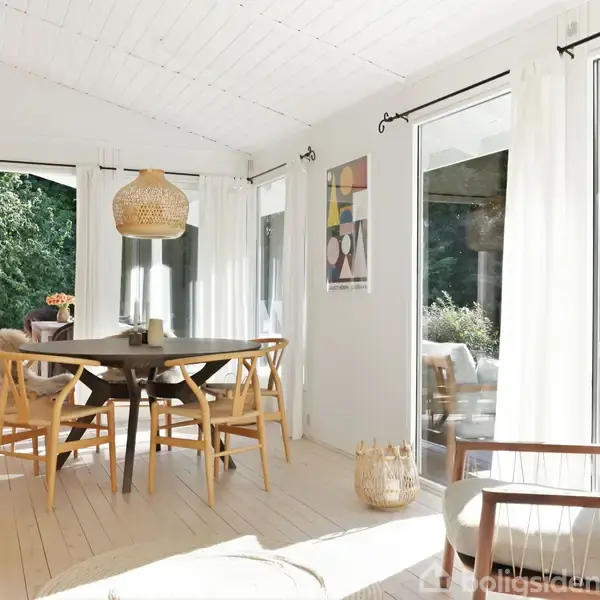 Spisebord med træstole står i et lyst rum med store vinduer, der åbner op til en grøn udsigt. Rundt om bordet hænger en flettet pendel, og væggene er hvide.