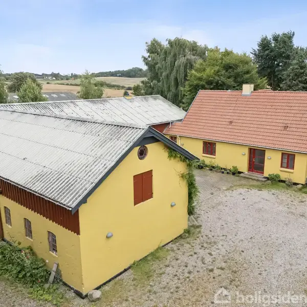 To gule bygninger med røde tage står ved siden af hinanden på en gårdsplads omgivet af træer og marker.
