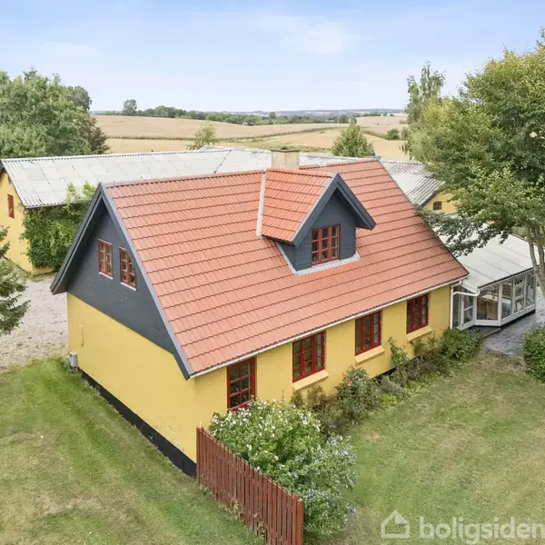 Gult hus med rødt tegltag og røde vinduer står omgivet af grønt græs og træer. Ved siden af huset ses en glasudbygning og et mindre gult bygning.
