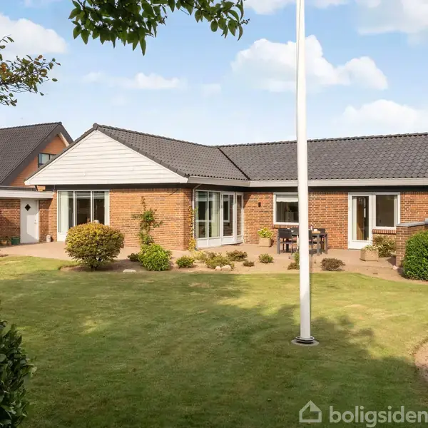 Et enfamiliehus i mursten med sorte tagsten står i en velplejet have. Flagstang i forgrunden og hække med grønne buske rundt om. Himmel med lette skyer.