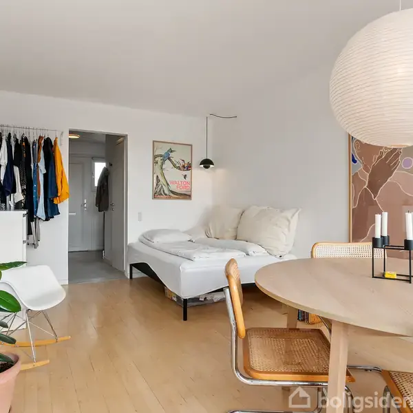 Soveværelse indeholder en seng med puder, omgivet af en moderne ramme. Rundt om er et spisebord med stole, en grøn plante i hjørnet, og en åben dør ind til garderobeområde.