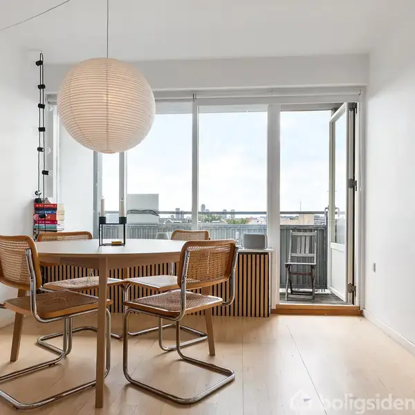 Et rundt spisebord med fire stole står i et lyst rum. En stor papirlampe hænger over bordet. Rummet har store vinduer og en altandør med udsigt. En plante er i hjørnet.