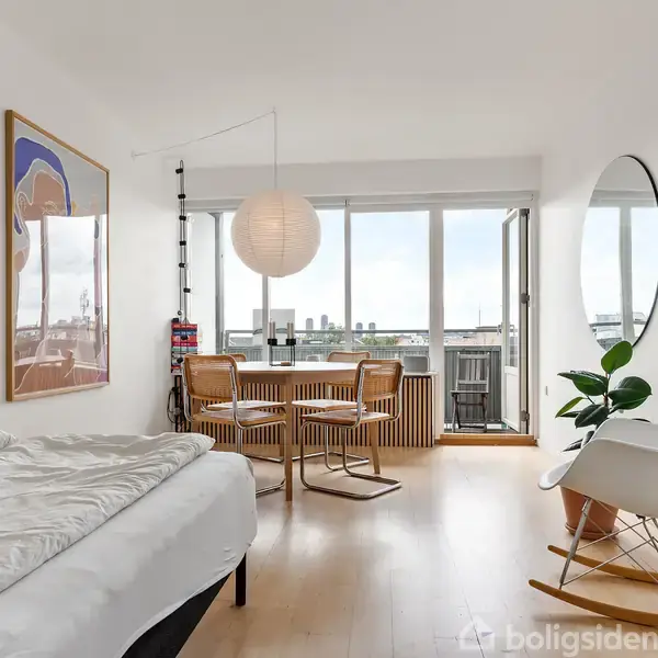 Soveværelse har en seng med hvidt sengetøj, en rund lampe over et spisebord med stole, en kommode og en stor plante. Store vinduer giver udsigt over byen.