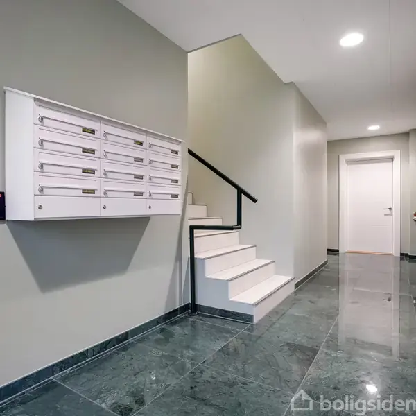 Postkasser hænger på en lys grøn væg i en bygning. Trapperne ved siden af fører opad. Rummet har en skinnende, mørk flisegulvbelægning og hvide døre i baggrunden.