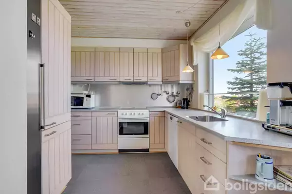 Et lyst køkken med træskabe står langs væggene. Komfuret og mikrobølgeovnen er centralt placeret. Fra det store vindue ses en udsigt med et træ udenfor.