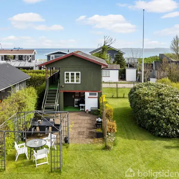Et træhus med en trappe fører op til en terrasse; omgivet af en velplejet have med et spisearrangement under en pergola. I baggrunden ses havudsigten.