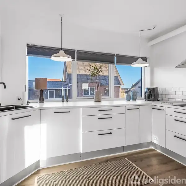 Hvidt, moderne køkken med hvide skabe og skuffer, sort bordplade. Indbygget ovn til venstre, rustfri emhætte over komfur til højre. Stort vindue med udsigt til huse udenfor.