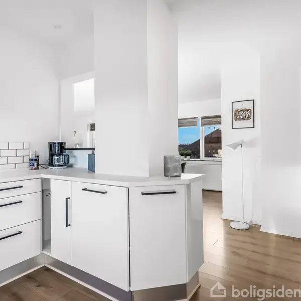 Det moderne køkken har hvide skabe og en stænkkant af hvide fliser. En emhætte hænger over komfuret. I baggrunden ses en lys stue med trægulve.