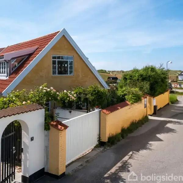 En gul villa med rødt tag står bag en hvid port omgivet af grønne planter. Villaen er placeret på en lille vej tæt på kysten med en blå himmel i baggrunden.