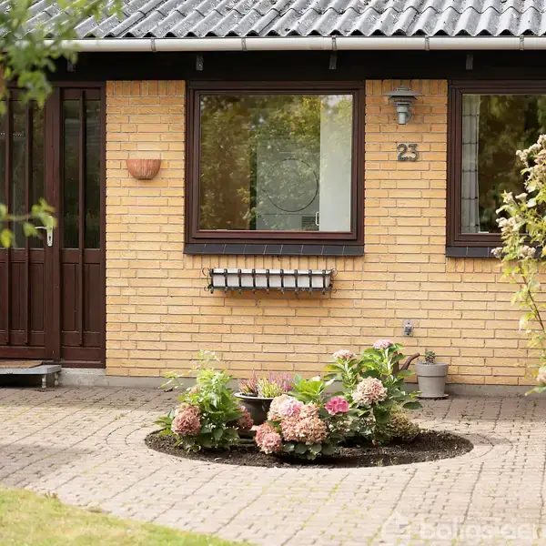 Gult murstenshus har hoveddør og store vinduer. Foran er en cirkulær blomsterbed med pink blomster. Husnummeret "23" er på væggen. Omkringliggende haver og belægning skaber en indbydende atmosfære.