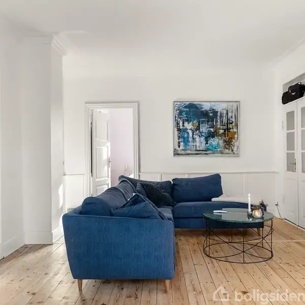 En blå sofa står stille i en stue med trægulv. Væggene er hvide, og der hænger et farverigt maleri over sofaen. En fladskærm og et rundt sofabord er også synlige.