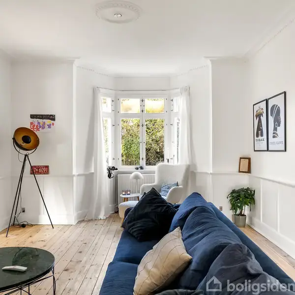 Sofaen står i en stue med hvidmalede vægge, parketgulv og et stort vindue. En plante og vægkunst dekorerer rummet. Træbord med stearinlys og flaske ved siden af.